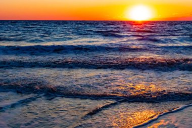 Güneşin ufukta battığı nefes kesici bir okyanus gün batımı dalgalanan mavi suya sıcak bir altın parıltı saçıyor. Huzurlu ve resimli sahil manzarası.