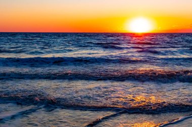 Güneşin ufukta battığı nefes kesici bir okyanus gün batımı dalgalanan mavi suya sıcak bir altın parıltı saçıyor. Huzurlu ve resimli sahil manzarası.