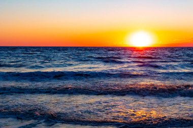 Güneşin ufukta battığı nefes kesici bir okyanus gün batımı dalgalanan mavi suya sıcak bir altın parıltı saçıyor. Huzurlu ve resimli sahil manzarası.