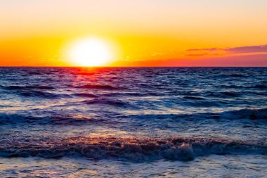Güneşin ufukta battığı nefes kesici bir okyanus gün batımı dalgalanan mavi suya sıcak bir altın parıltı saçıyor. Huzurlu ve resimli sahil manzarası.