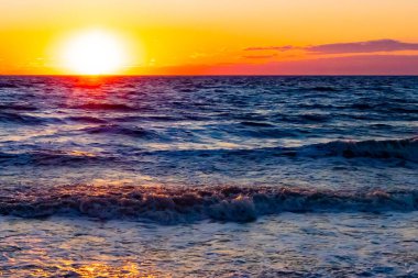 Güneşin ufukta battığı nefes kesici bir okyanus gün batımı dalgalanan mavi suya sıcak bir altın parıltı saçıyor. Huzurlu ve resimli sahil manzarası.