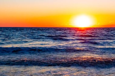 Güneşin ufukta battığı nefes kesici bir okyanus gün batımı dalgalanan mavi suya sıcak bir altın parıltı saçıyor. Huzurlu ve resimli sahil manzarası.