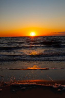 Güneşin ufukta battığı nefes kesici bir okyanus gün batımı dalgalanan mavi suya sıcak bir altın parıltı saçıyor. Huzurlu ve resimli sahil manzarası.