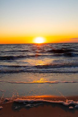 Güneşin ufukta battığı nefes kesici bir okyanus gün batımı dalgalanan mavi suya sıcak bir altın parıltı saçıyor. Huzurlu ve resimli sahil manzarası.
