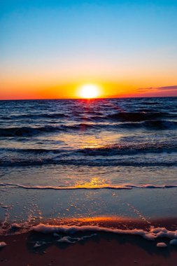 Güneşin ufukta battığı nefes kesici bir okyanus gün batımı dalgalanan mavi suya sıcak bir altın parıltı saçıyor. Huzurlu ve resimli sahil manzarası.
