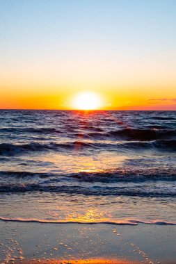 Güneşin ufukta battığı nefes kesici bir okyanus gün batımı dalgalanan mavi suya sıcak bir altın parıltı saçıyor. Huzurlu ve resimli sahil manzarası.