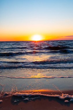 Güneşin ufukta battığı nefes kesici bir okyanus gün batımı dalgalanan mavi suya sıcak bir altın parıltı saçıyor. Huzurlu ve resimli sahil manzarası.