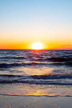Güneşin ufukta battığı nefes kesici bir okyanus gün batımı dalgalanan mavi suya sıcak bir altın parıltı saçıyor. Huzurlu ve resimli sahil manzarası.