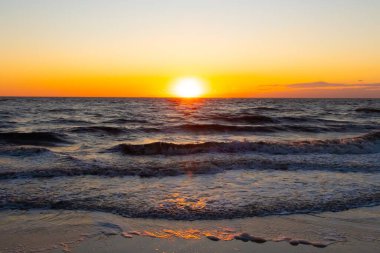 Güneşin ufukta battığı nefes kesici bir okyanus gün batımı dalgalanan mavi suya sıcak bir altın parıltı saçıyor. Huzurlu ve resimli sahil manzarası.