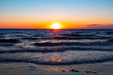 Güneşin ufukta battığı nefes kesici bir okyanus gün batımı dalgalanan mavi suya sıcak bir altın parıltı saçıyor. Huzurlu ve resimli sahil manzarası.