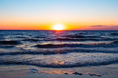Güneşin ufukta battığı nefes kesici bir okyanus gün batımı dalgalanan mavi suya sıcak bir altın parıltı saçıyor. Huzurlu ve resimli sahil manzarası.
