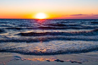 Güneşin ufukta battığı nefes kesici bir okyanus gün batımı dalgalanan mavi suya sıcak bir altın parıltı saçıyor. Huzurlu ve resimli sahil manzarası.