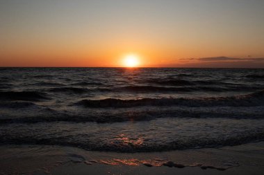 Güneşin ufukta battığı nefes kesici bir okyanus gün batımı dalgalanan mavi suya sıcak bir altın parıltı saçıyor. Huzurlu ve resimli sahil manzarası.