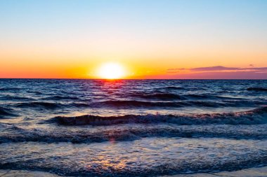 Güneşin ufukta battığı nefes kesici bir okyanus gün batımı dalgalanan mavi suya sıcak bir altın parıltı saçıyor. Huzurlu ve resimli sahil manzarası.