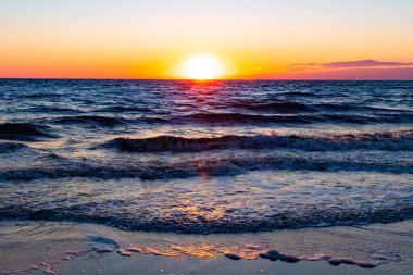 Güneşin ufukta battığı nefes kesici bir okyanus gün batımı dalgalanan mavi suya sıcak bir altın parıltı saçıyor. Huzurlu ve resimli sahil manzarası.