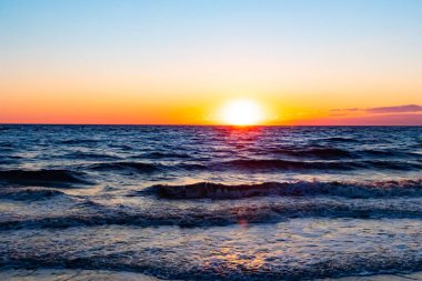 Güneşin ufukta battığı nefes kesici bir okyanus gün batımı dalgalanan mavi suya sıcak bir altın parıltı saçıyor. Huzurlu ve resimli sahil manzarası.