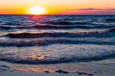 Güneşin ufukta battığı nefes kesici bir okyanus gün batımı dalgalanan mavi suya sıcak bir altın parıltı saçıyor. Huzurlu ve resimli sahil manzarası.