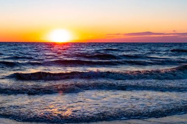 Güneşin ufukta battığı nefes kesici bir okyanus gün batımı dalgalanan mavi suya sıcak bir altın parıltı saçıyor. Huzurlu ve resimli sahil manzarası.