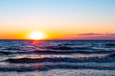 Güneşin ufukta battığı nefes kesici bir okyanus gün batımı dalgalanan mavi suya sıcak bir altın parıltı saçıyor. Huzurlu ve resimli sahil manzarası.