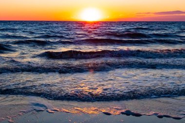 Güneşin ufukta battığı nefes kesici bir okyanus gün batımı dalgalanan mavi suya sıcak bir altın parıltı saçıyor. Huzurlu ve resimli sahil manzarası.