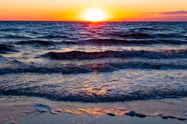 Güneşin ufukta battığı nefes kesici bir okyanus gün batımı dalgalanan mavi suya sıcak bir altın parıltı saçıyor. Huzurlu ve resimli sahil manzarası.