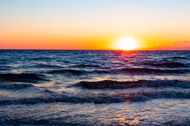 Güneşin ufukta battığı nefes kesici bir okyanus gün batımı dalgalanan mavi suya sıcak bir altın parıltı saçıyor. Huzurlu ve resimli sahil manzarası.
