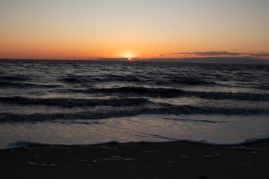 Güneşin ufukta battığı nefes kesici bir okyanus gün batımı dalgalanan mavi suya sıcak bir altın parıltı saçıyor. Huzurlu ve resimli sahil manzarası.