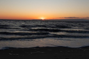 Güneşin ufukta battığı nefes kesici bir okyanus gün batımı dalgalanan mavi suya sıcak bir altın parıltı saçıyor. Huzurlu ve resimli sahil manzarası.