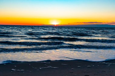 Güneşin ufukta battığı nefes kesici bir okyanus gün batımı dalgalanan mavi suya sıcak bir altın parıltı saçıyor. Huzurlu ve resimli sahil manzarası.