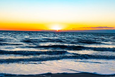 Güneşin ufukta battığı nefes kesici bir okyanus gün batımı dalgalanan mavi suya sıcak bir altın parıltı saçıyor. Huzurlu ve resimli sahil manzarası.