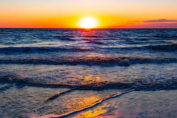 Güneşin ufukta battığı nefes kesici bir okyanus gün batımı dalgalanan mavi suya sıcak bir altın parıltı saçıyor. Huzurlu ve resimli sahil manzarası.