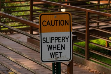 Parkta ıslandığında kaygan olduğuna dair bir işaret. Arkasında kırmızı bir köprü var. Paslı metal yıpranmış yaşını gösteriyor..