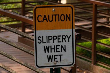 Parkta ıslandığında kaygan olduğuna dair bir işaret. Arkasında kırmızı bir köprü var. Paslı metal yıpranmış yaşını gösteriyor..