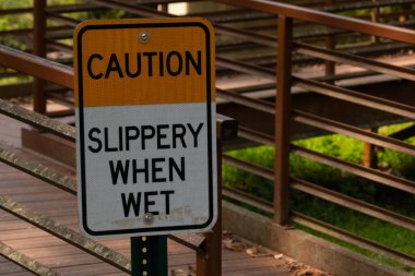 Parkta ıslandığında kaygan olduğuna dair bir işaret. Arkasında kırmızı bir köprü var. Paslı metal yıpranmış yaşını gösteriyor..