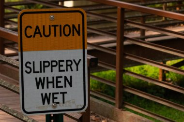 Parkta ıslandığında kaygan olduğuna dair bir işaret. Arkasında kırmızı bir köprü var. Paslı metal yıpranmış yaşını gösteriyor..