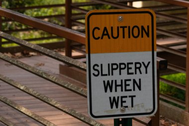 Parkta ıslandığında kaygan olduğuna dair bir işaret. Arkasında kırmızı bir köprü var. Paslı metal yıpranmış yaşını gösteriyor..