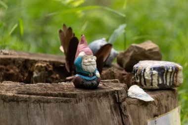 Kelebek kanatlı tuhaf bir bahçe cücesi heykeli, kayalar ve yeşilliklerle çevrili bir ağaç kütüğünün tepesinde oturur ve doğa ya da fantezi temalar için mükemmel büyüleyici ve eğlenceli bir açık hava sahnesi yaratır.