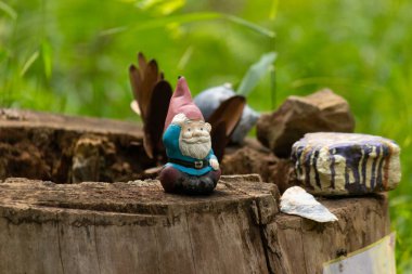 Kelebek kanatlı tuhaf bir bahçe cücesi heykeli, kayalar ve yeşilliklerle çevrili bir ağaç kütüğünün tepesinde oturur ve doğa ya da fantezi temalar için mükemmel büyüleyici ve eğlenceli bir açık hava sahnesi yaratır.