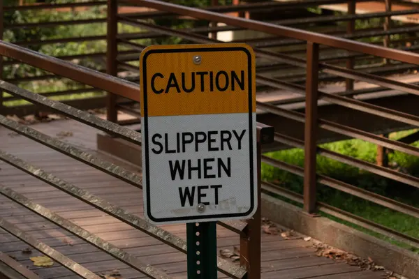 Parkta ıslandığında kaygan olduğuna dair bir işaret. Arkasında kırmızı bir köprü var. Paslı metal yıpranmış yaşını gösteriyor..