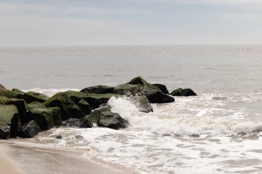 Bu, suya uzanan bir iskelenin resmi. Dalgalar etrafa köpük saçıyor. Körfezden çıkan büyük siyah kayaların üzerinde yeşil yosun yetişiyor..