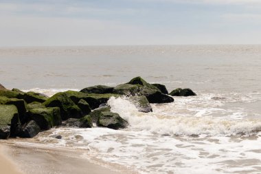 Bu, suya uzanan bir iskelenin resmi. Dalgalar etrafa köpük saçıyor. Körfezden çıkan büyük siyah kayaların üzerinde yeşil yosun yetişiyor..