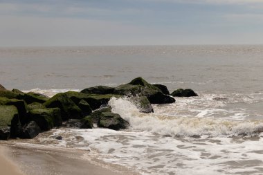 Bu, suya uzanan bir iskelenin resmi. Dalgalar etrafa köpük saçıyor. Körfezden çıkan büyük siyah kayaların üzerinde yeşil yosun yetişiyor..