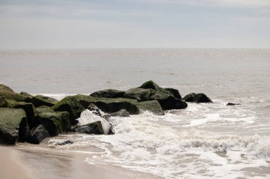 Bu, suya uzanan bir iskelenin resmi. Dalgalar etrafa köpük saçıyor. Körfezden çıkan büyük siyah kayaların üzerinde yeşil yosun yetişiyor..