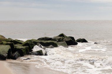 Bu, suya uzanan bir iskelenin resmi. Dalgalar etrafa köpük saçıyor. Körfezden çıkan büyük siyah kayaların üzerinde yeşil yosun yetişiyor..