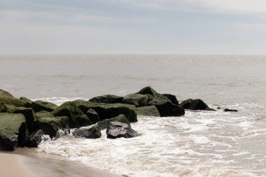 Bu, suya uzanan bir iskelenin resmi. Dalgalar etrafa köpük saçıyor. Körfezden çıkan büyük siyah kayaların üzerinde yeşil yosun yetişiyor..