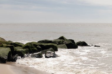Bu, suya uzanan bir iskelenin resmi. Dalgalar etrafa köpük saçıyor. Körfezden çıkan büyük siyah kayaların üzerinde yeşil yosun yetişiyor..