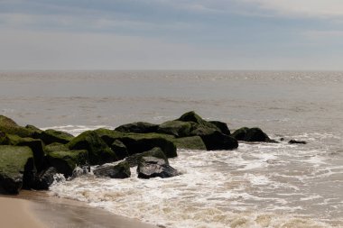 Bu, suya uzanan bir iskelenin resmi. Dalgalar etrafa köpük saçıyor. Körfezden çıkan büyük siyah kayaların üzerinde yeşil yosun yetişiyor..
