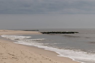 Jersey sahilinin güzel bir görüntüsü. Dalgaların beyaz köpüğü kumsalın güzel kumlarına çarpıyor. Siyah taşlı küçük bir iskele görülebilir. Mavi gökyüzünde beyaz bulutlar var..