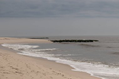 Jersey sahilinin güzel bir görüntüsü. Dalgaların beyaz köpüğü kumsalın güzel kumlarına çarpıyor. Siyah taşlı küçük bir iskele görülebilir. Mavi gökyüzünde beyaz bulutlar var..