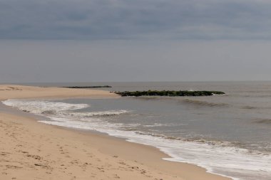 Jersey sahilinin güzel bir görüntüsü. Dalgaların beyaz köpüğü kumsalın güzel kumlarına çarpıyor. Siyah taşlı küçük bir iskele görülebilir. Mavi gökyüzünde beyaz bulutlar var..